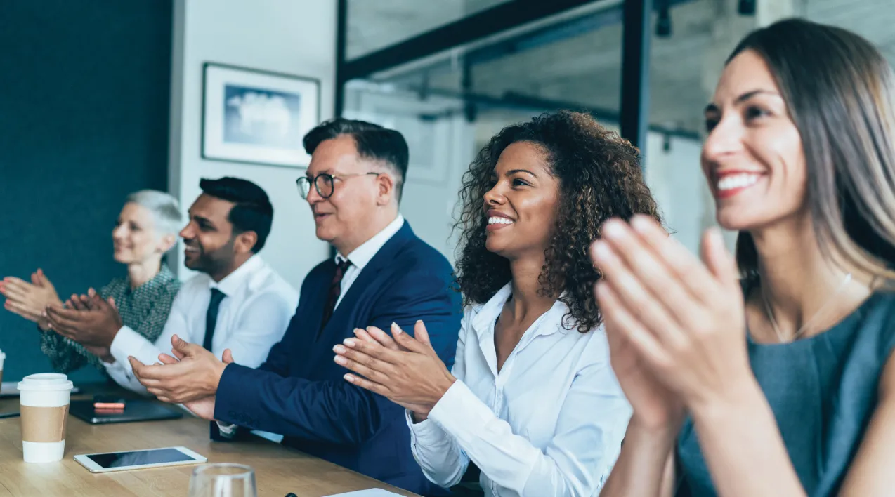 A group of coworkers sit around a conference room table clapping.