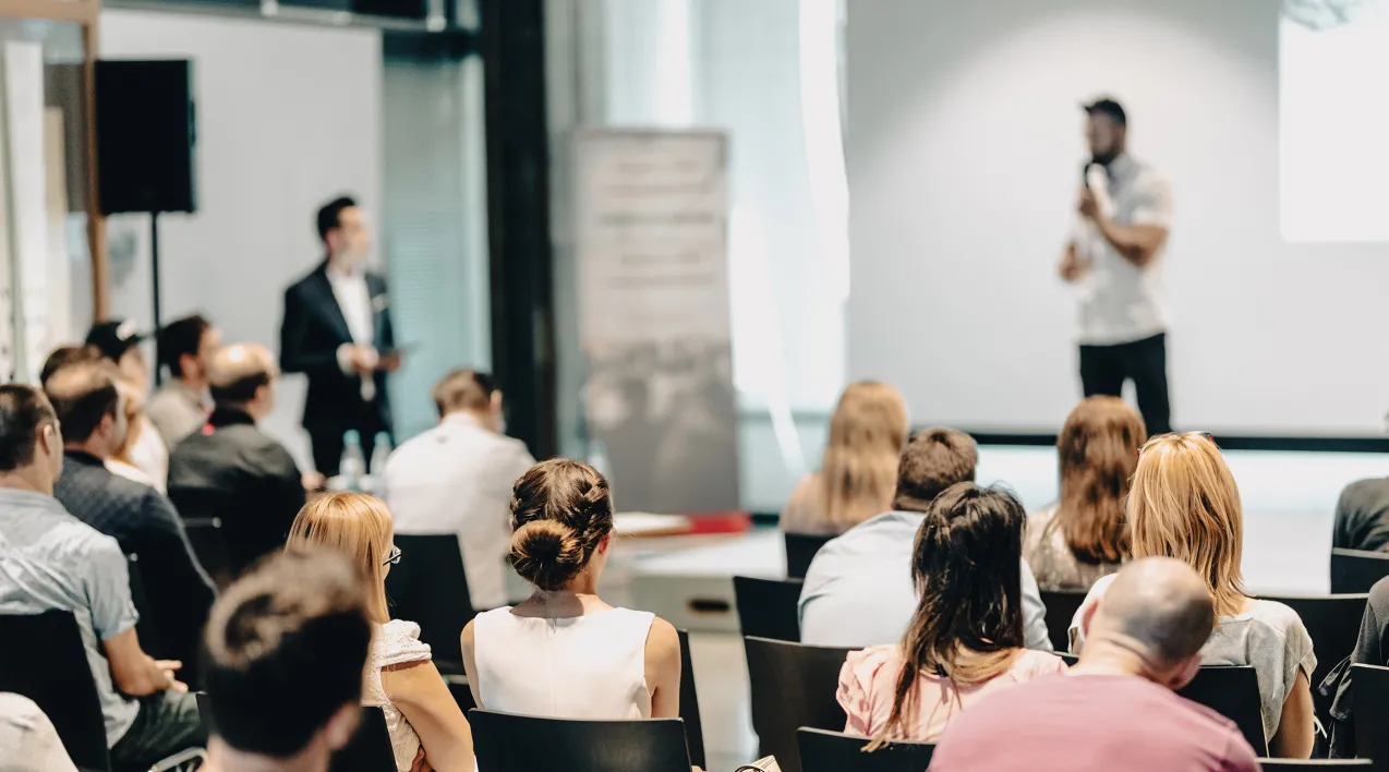 A group at a conference sits around a stage while man presents.