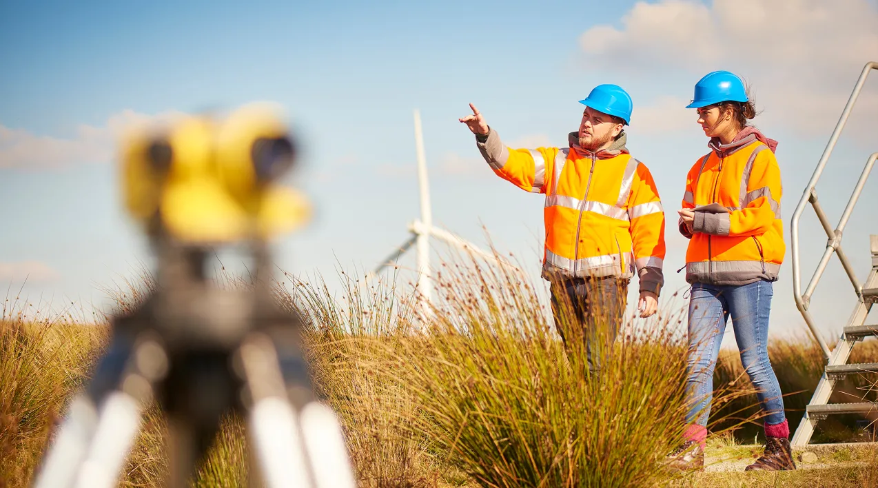 A man and a woman in high-visibility gear work on a site with a wind mill in the background.