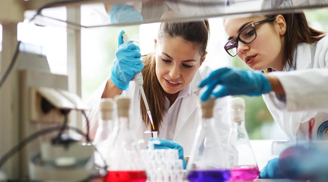 Two women work on a lab counter using a pipette and multiple colored liquids in flasks.