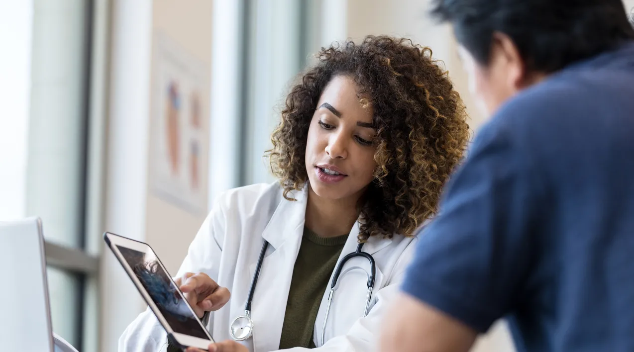 A healthcare provider speaks with a patient while looking at a tablet.